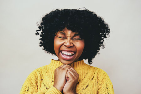 Closeup portrait of cheerful Black woman laughing with eyes closed holding hands wearing yellow sweater isolated against gray backgroundの写真素材
