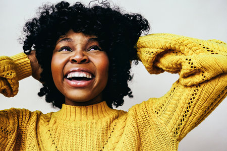 A cheerful moment. Closeup portrait of smiling black woman with hands on curly hair looking up wearing a yellow sweater isolated against a gray backgroundの写真素材