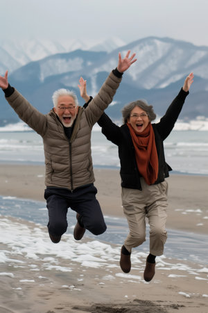 A couple of older people are jumping in the air on a beach. They are both smiling and seem to be enjoying themselvesの素材