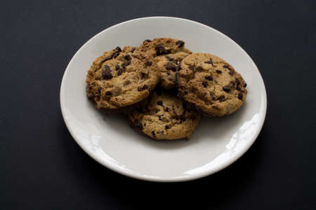 Four chocolate chip cookies on white plate on black background.