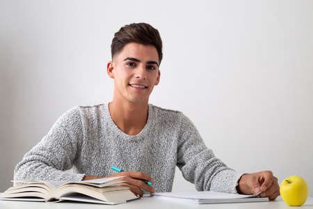 smiling teenager studying on a desk. On the table there is a book, papers and a pen. A cup and an apple too. He look like happyの写真素材