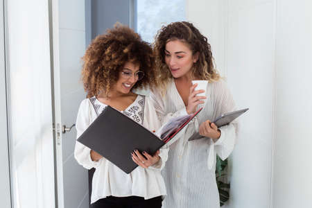 Two young women coworkers speaking and working in the office. They are smiling and happy. They are...