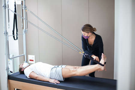young man wearing mask doing pilates exercise on reformer with his female instructor during covid-19の写真素材