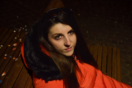 Young woman in orange jacket and sunglasses sitting on bench at night.の写真素材