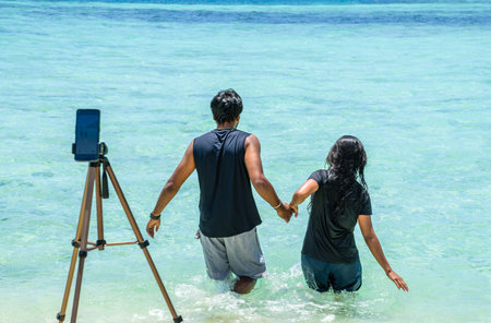 Indian couple taking pictures and videos with a tripod while walking towards the sea.の写真素材
