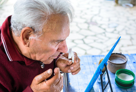 Old man with red sweater and gray hair shaving while watching his face on mirrorの写真素材