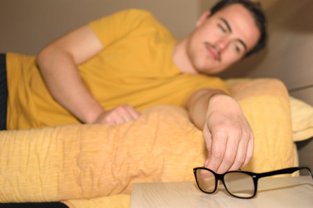 Young man holding his glasses as he gets out of bed with dim light