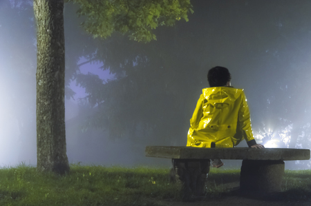 Woman with yellow raincoat sitting on a stone bank on a hill a foggy night in autumn の写真素材