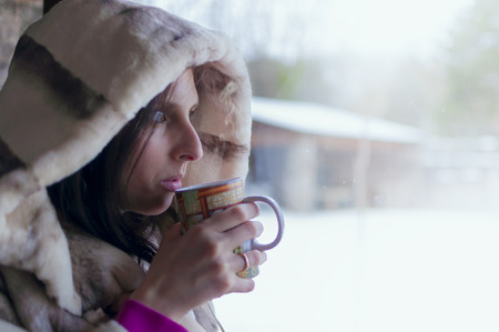 Woman drinking tea in the window watching the cold winter day meaning through the window wrapped in a fur coat - 4の写真素材