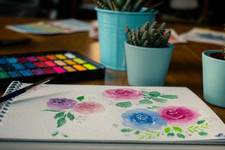 Close-up watercolor roses painting surrounded by three cactuses and a watercolor palette on a wooden table from a frontal viewの写真素材
