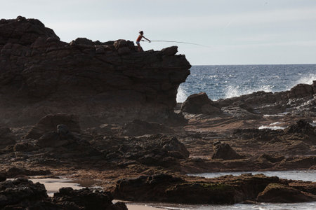 fisherman silhouette on rocks on sandy beach at sunsetの写真素材