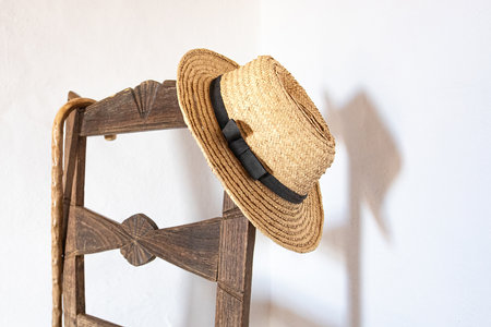 Straw hat hanging on a wooden chair against a white wall.の写真素材