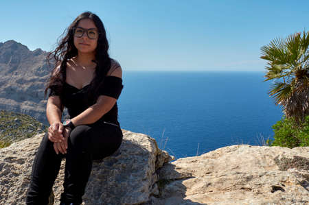 Latin girl, with long wavy brunette hair, posing for, portrait, on a spring day with glasses, sitting on the cliff of Cap de Formentor Mallorca, Balearic islands, Spain,の写真素材