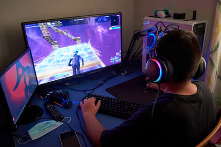 Back view of teenage gamer boy playing video games online on computer in dark room, wearing headphones with microphone and using backlit colorful keyboardの写真素材