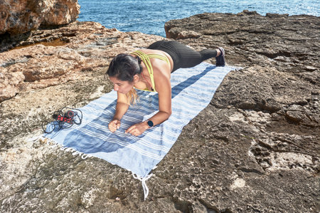 FITNESS WOMAN IN SPORTS SET TRAINING WITH ELASTIC BAND, WEIGHTS, GYM EXERCISES, IN FRONT OF THE WATER.の写真素材
