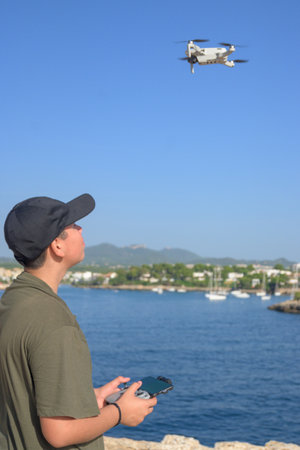 happy teen boy, flying drone on mediterranean coast, against blue sky during sunny day Spain,...