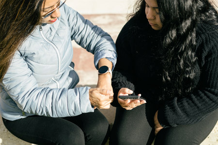 Hispanic mother in sunglasses checking time on smartwatch while sitting on bench with daughter using smartphone in park on sunny dayの写真素材