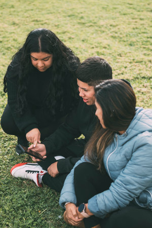 three people of Hispanic-Latino ethnicity, sitting on the ground in the park with smart devicesの写真素材