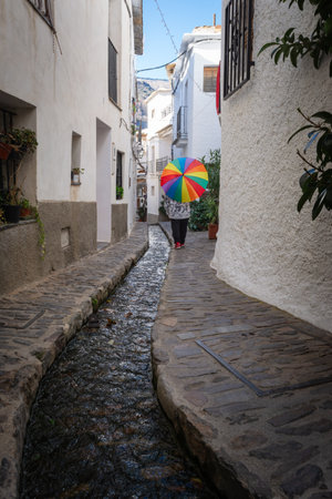 Person with a colorful umbrella walking along a narrow stone path with a stream in a white building alley, Alpujarras, granada, andalucia, espa aの写真素材