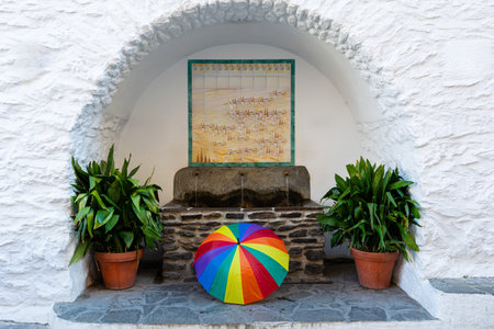 A colorful rainbow umbrella placed in front of an arch with white walls, potted plants, and hanging artwork, Alpujarras, granada, andalucia, espa aの写真素材