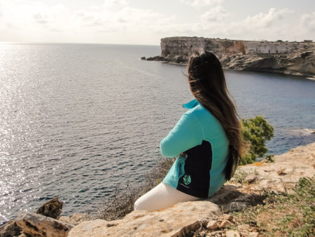 Woman sitting on a cliff gazing at the ocean under a sunny sky, looking out over the mediterranean sea cliffs, in Mallorca Balearic Islands Spainの写真素材