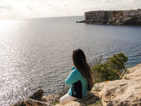 Woman perched on a cliff edge, viewing the expansive ocean under the sun, latin woman looking out over the mediterranean sea cliffs, in mallorca balearic islands spainの写真素材