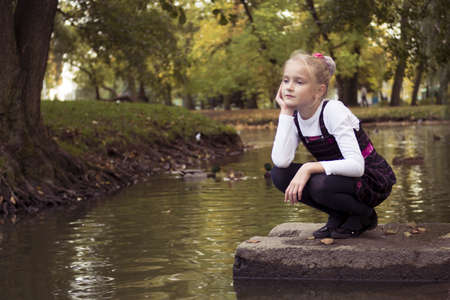 Little preteen girl sitting near the lake with ducks in park at autumnの写真素材
