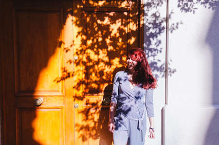 A young adult redhead female smiling in front of a wood entrance door in the street with shadows of...