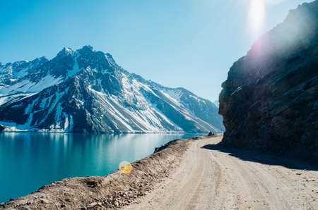 A dramatic panorama of El Yeso Dam with turquoise colour and sun flares reflecting in the water in the Andes Mountains with snow on the peaks and rock parts of the mountain exposed and a dirt road.の写真素材