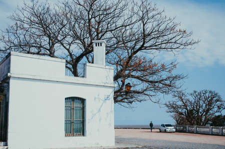 Colonia Del Sacramento, Uruguay - September 10, 2018: A medium sized colonial house with white walls in front of an old tree without leaves and a small male figure in the streets in front of a car.のeditorial素材