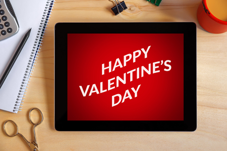 Valentine's day concept on tablet screen with office objects on wooden desk. All screen content is designed by me. View from above.の写真素材