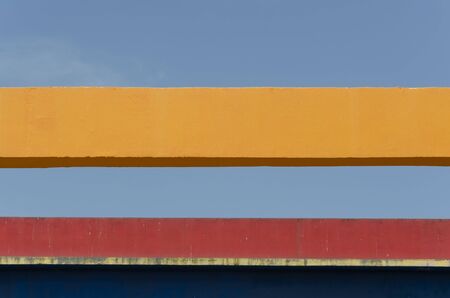 Urban detail, a blue and red wall with an eave that casts shadow, a yellow girder and a clear sky,...