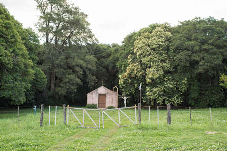 Route 21, Soriano / Uruguay; Dec 26, 2018: Small construction with a cross on top and a crucified Christ next to it, in a rural setting. A worn-out flag of Uruguay waves at the gate.のeditorial素材