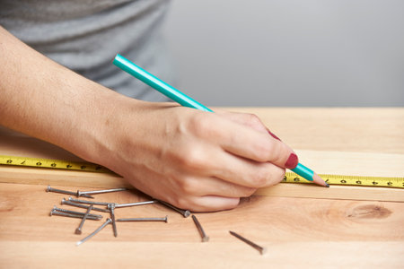 Detail of a young woman working in carpentry, marking measurements on a wooden strip with a pencil.