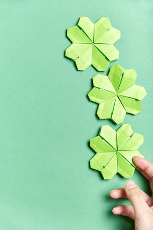 Origami four-leaf shamrocks, made of light green paper, on green background. A young hand is placing one of the clovers at the bottom. Concepts of luck and St. Patrick's Day. Image with copy space.の写真素材