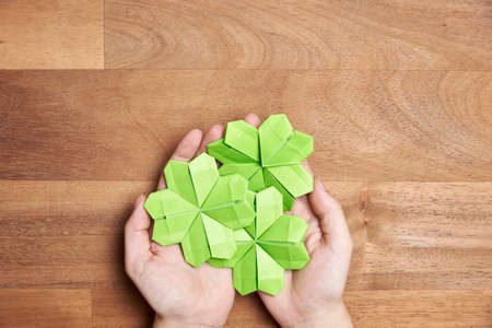 The hands of a young woman hold three origami clovers made of light green paper on a wooden background. Concepts of luck and St. Patrick's Day. Image with copy space.の写真素材