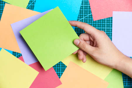Young female hand holding a square of blank green paper over other colored papers and a cutting mat....
