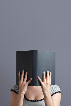 Young woman with her face hidden behind a black book that she holds with both hands, open, reading...