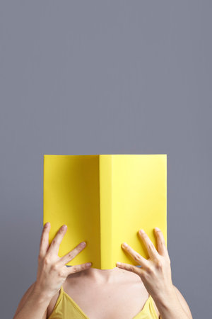 Young woman with her face hidden behind a yellow book that she holds with both hands, open, reading it. Portrait with gray background and copy space. Concept of reading.の写真素材