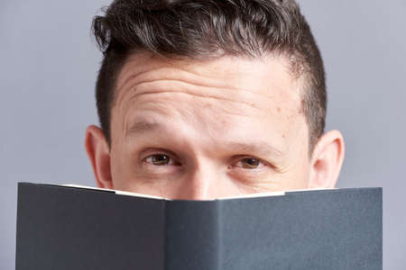 Young man looking at the camera raising an eyebrow, peeking out from behind an open black book. Frontal portrait with gray background.の写真素材