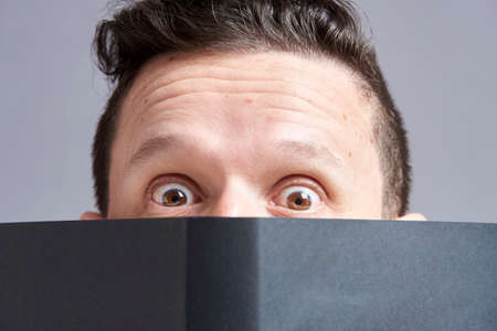 Young man staring in surprise at the camera, eyes bulging, peering out from behind an open black book. Frontal portrait with gray background.の写真素材