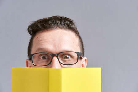 Young man, wearing glasses, looking at the camera, peeking out from behind an open yellow book....