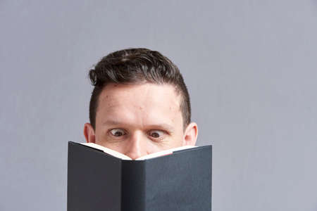 Young man reading a black book with squinted eyes; a comical, funny look. Frontal portrait with gray background and copy space.の写真素材
