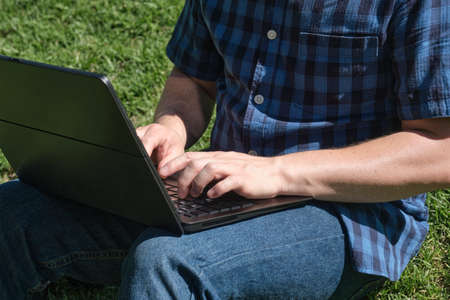 Unrecognizable young man typing on a laptop sitting on the grass in a park on a sunny day. Concept...