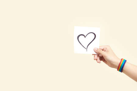 Young hand holding a paper with a hand-drawn heart, wearing a rainbow bracelet, symbol of the LGBT...