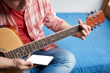 Young man learning to play guitar at home using his smart phone. Concepts of online learning education, hobbies and music.の写真素材