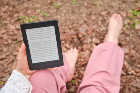Unrecognizable mature woman reading an ebook on an electronic reader sitting in a park. Concepts: technology and reading.の写真素材
