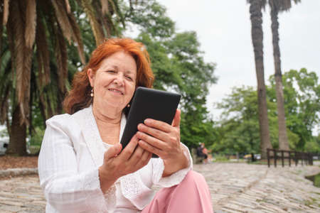 Mature hispanic woman reading an ebook on an electronic reader sitting in a park. Concepts: technology and reading. Image with copy spaceの写真素材