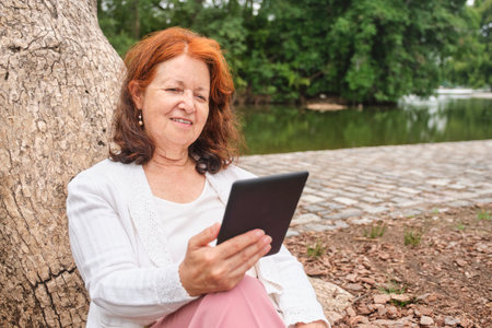 Mature hispanic woman reading an ebook on an electronic reader sitting in a park. Concepts:...