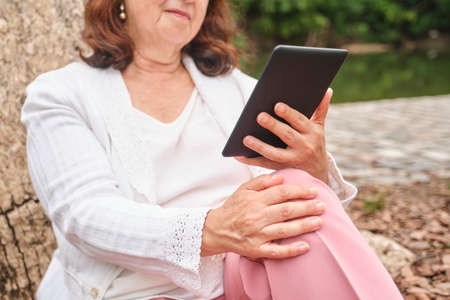 Unrecognizable mature woman reading an ebook on an electronic reader sitting by a lake in a park. Concepts: technology and reading.の写真素材